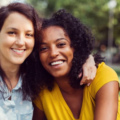 smiling white woman embracing and wrapping arm around smiling black woman