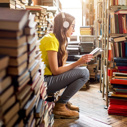 young woman wearing headphones surrounded by a heap of books in the library